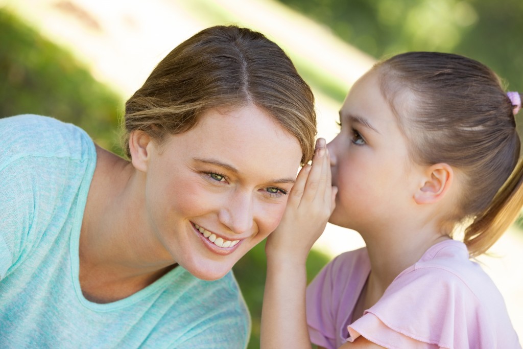 Close-up of a little young girl whispering secret into mother's ear at the park